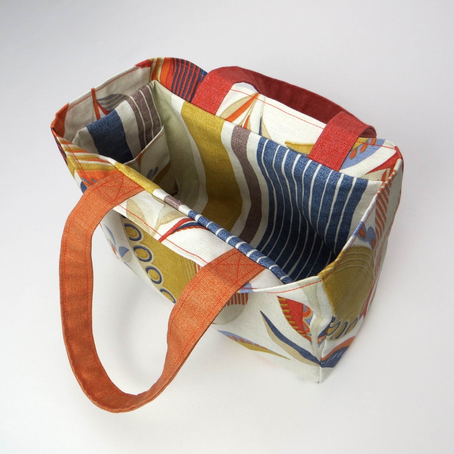 Small cotton tote bag with tulip pattern in shades of warm orange, rich ochre, and classic denim blue on an off-white background